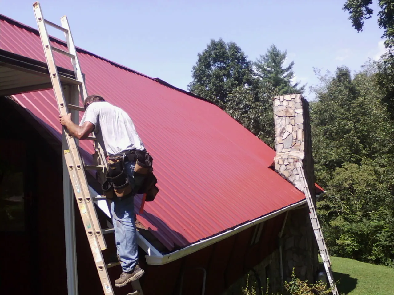 Roofer on a ladder beside a red metal roof and stone chimney — Soffit & Fascia Repair by Roofing Brothers in Pollocksville